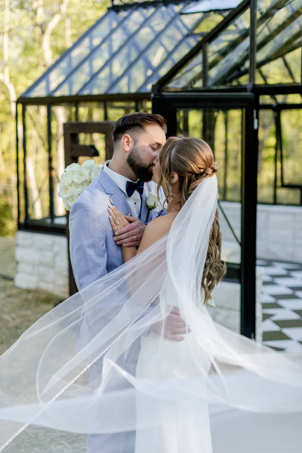 A couple shares a romantic kiss under a flowing veil in a scenic outdoor venue surrounded by greenery.