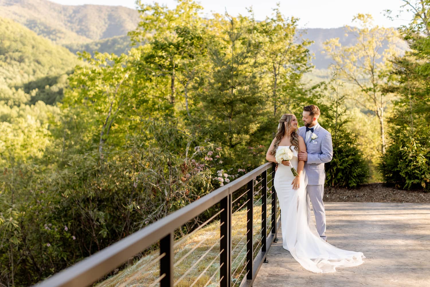 Couple in wedding attire stands on a balcony surrounded by lush greenery and mountains, celebrating their elopement.