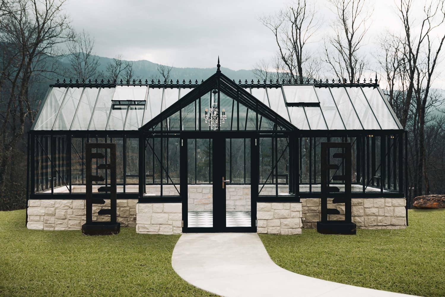 A European glass greenhouse with a stone foundation, surrounded by grassy areas and trees.
