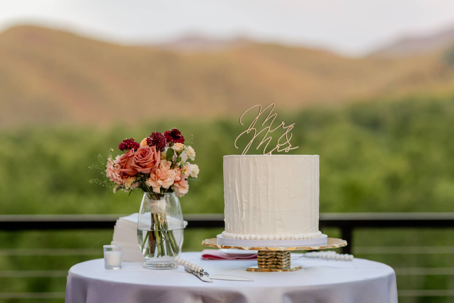 A wedding cake on a table beside a floral arrangement, with mountains in the background, perfect for elopements.