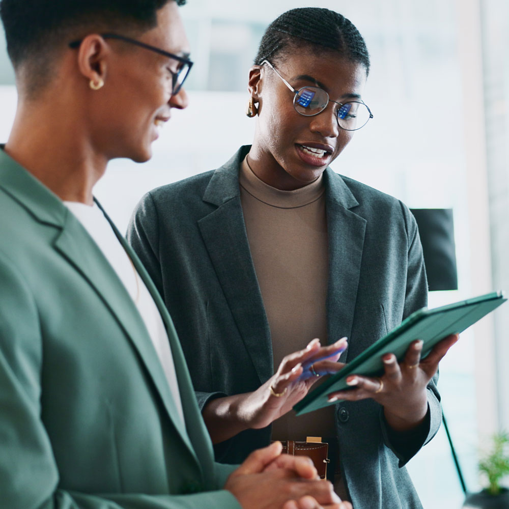 Two professionals in blazers discussing work while looking at a tablet in a modern office.