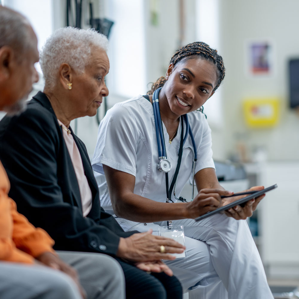 A female healthcare professional in white scrubs with a stethoscope talking to two elderly patients.