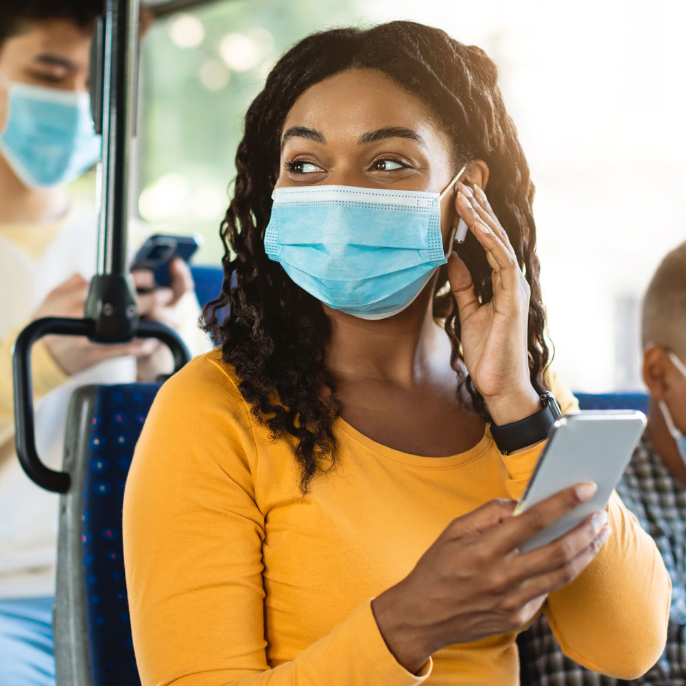 Woman wearing a blue face mask and yellow shirt holding a smartphone and adjusting an earbud on a bus.