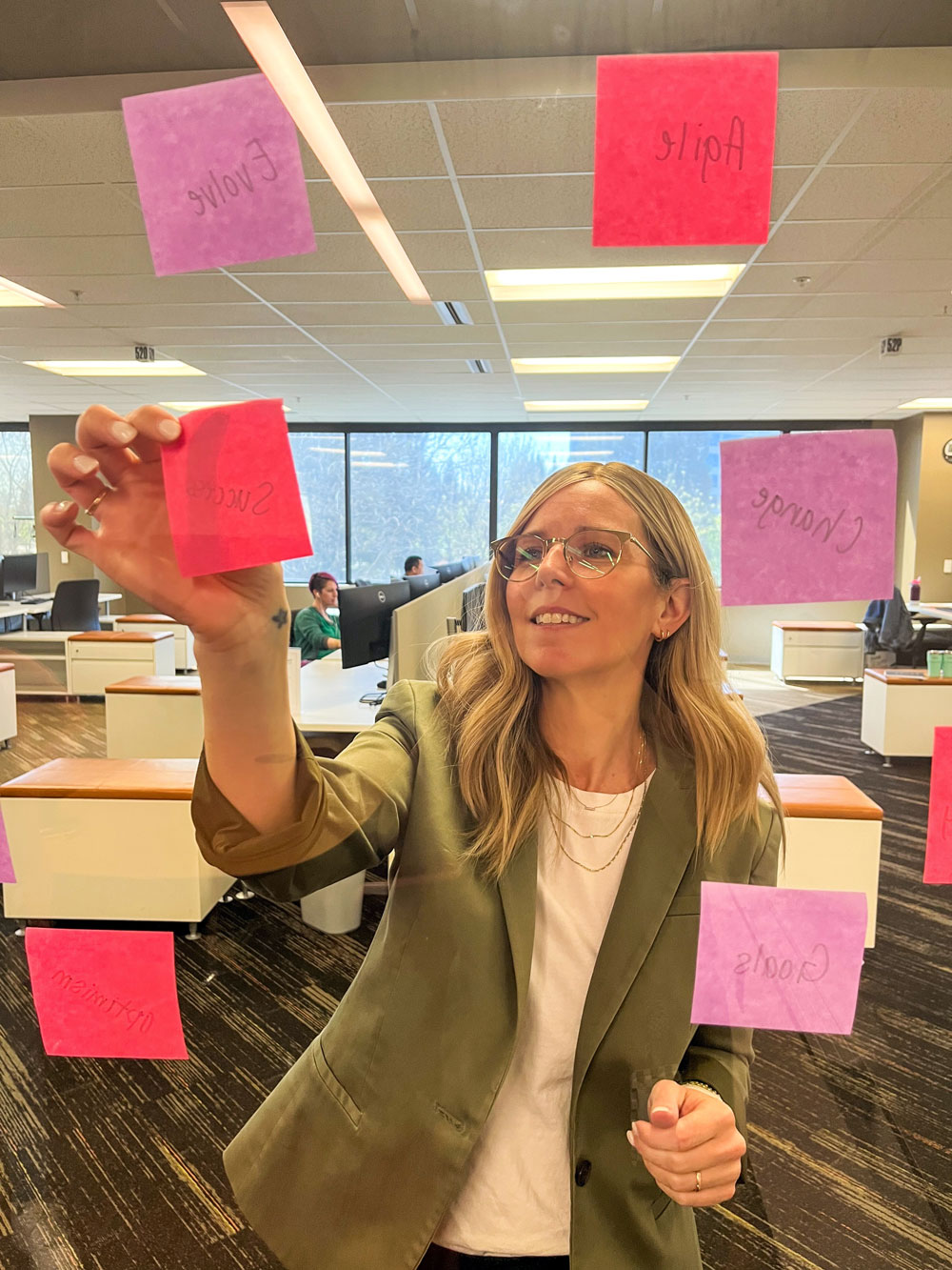 A woman in a green blazer writing on red and purple sticky notes attached to a glass wall in an office.