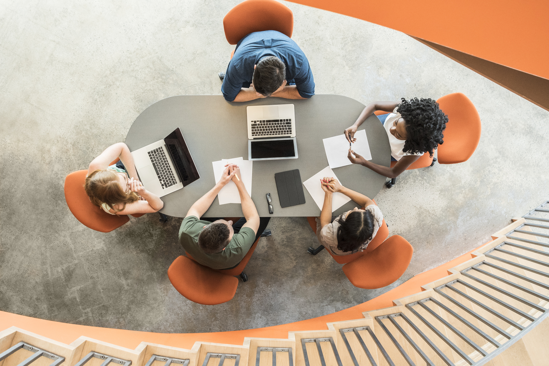 Overhead view of five diverse people seated around a table with laptops and papers, engaged in discussion in a modern space with orange chairs and curved staircase.