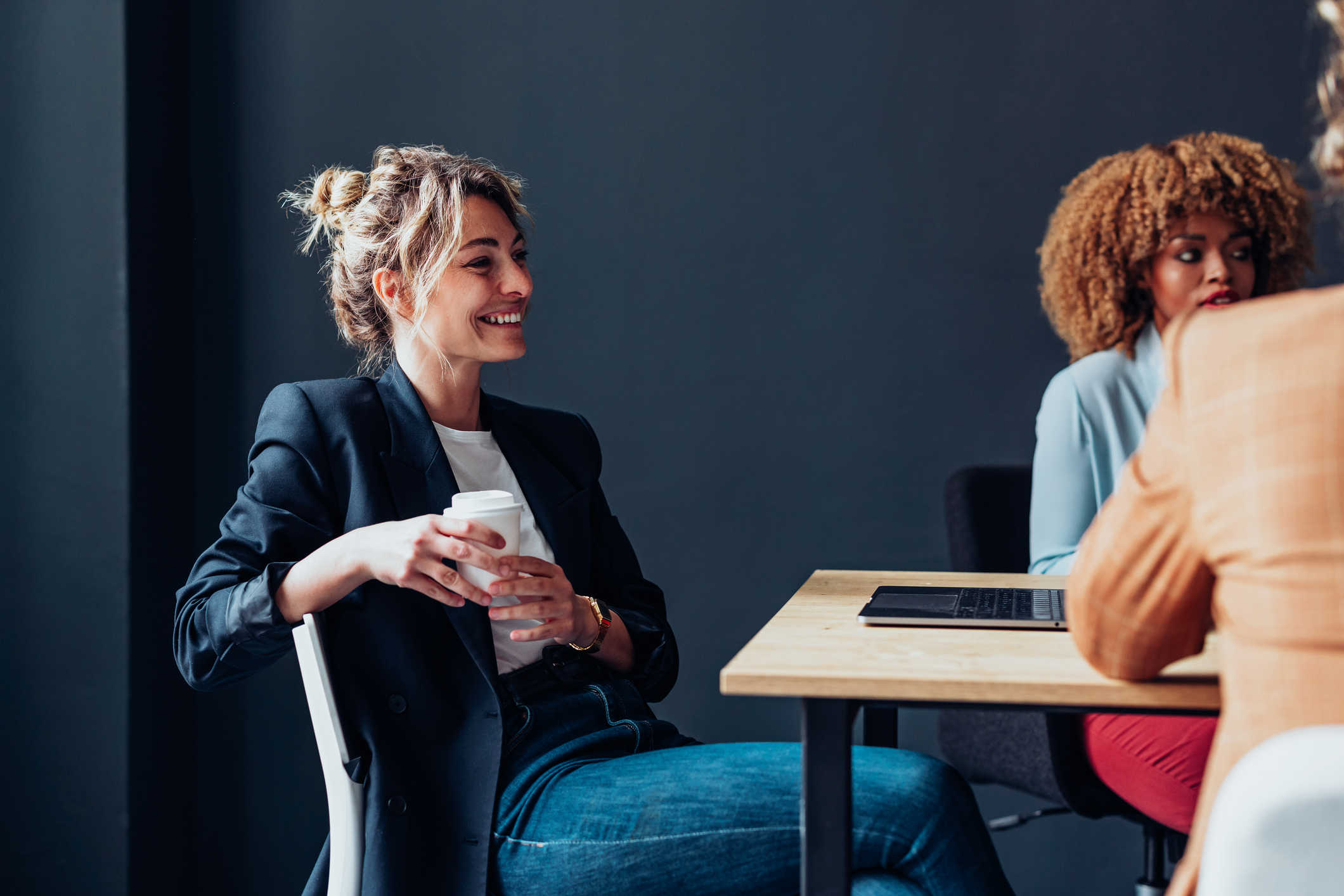Smiling woman in a black blazer holding a coffee cup seated at a table with two coworkers.