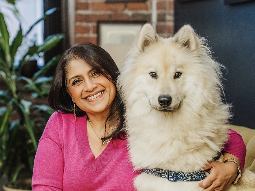 Mala Rafik, President and Co-Founder at MHJP, smiling warmly with long black hair, wearing a pink shirt, sitting with her dog, against an office background.