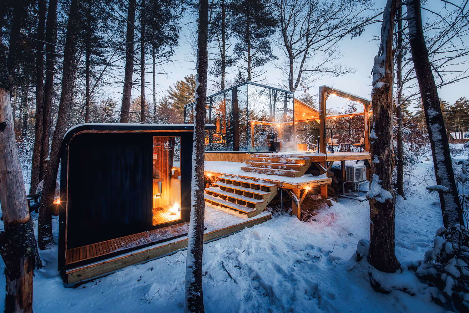 Warmly lit cabin exterior at sunset surrounded by snow and forest.