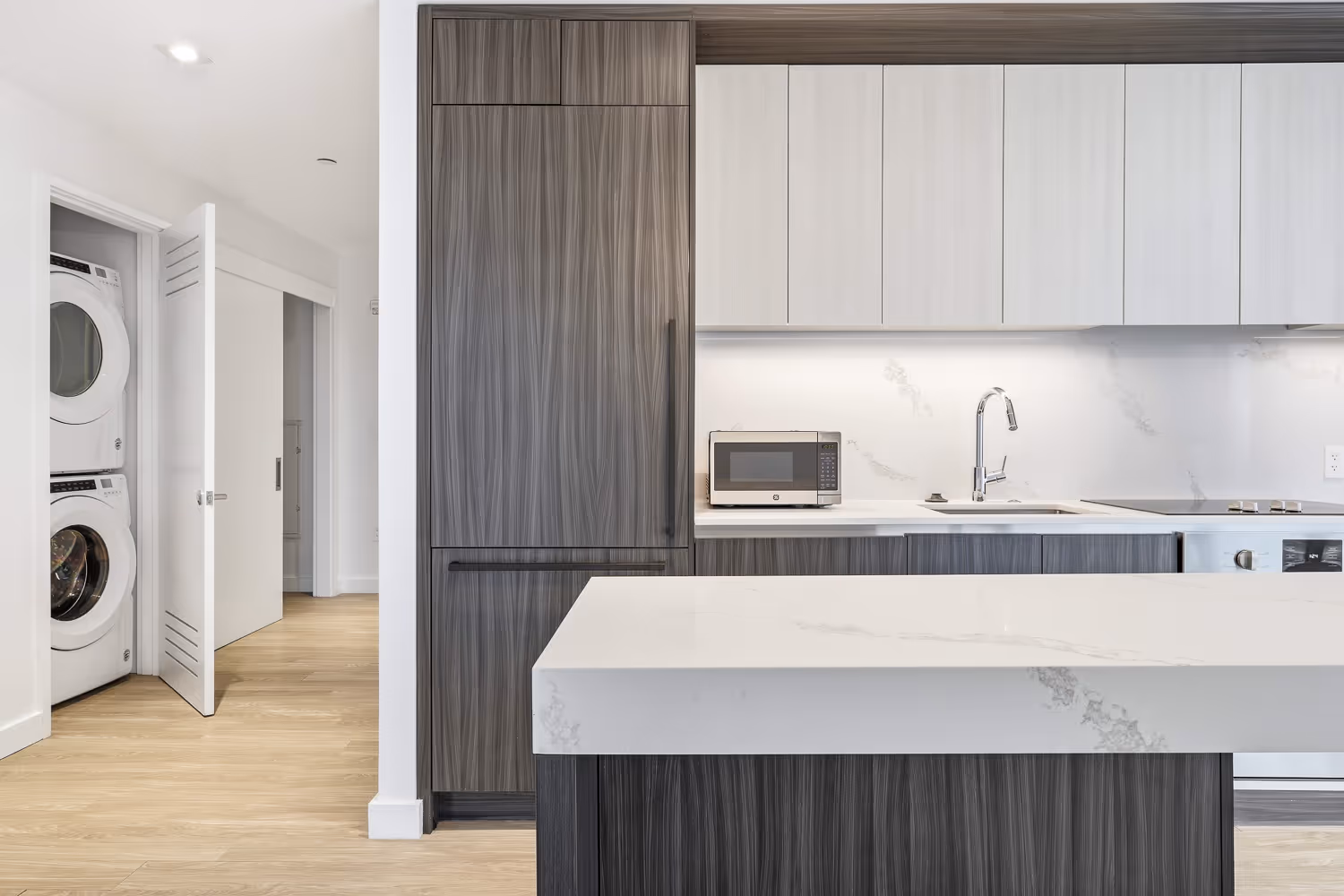 Modern kitchen with a white marble island, dark wood cabinets, a stainless steel microwave, sink with a chrome faucet, and stacked washer and dryer in an open closet.