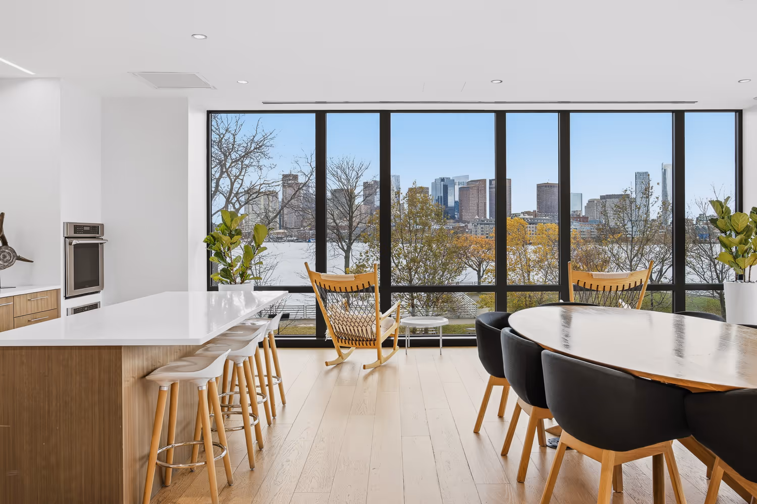 Modern dining and kitchen area with wooden stools, black chairs, rocking chairs, and large windows overlooking a river and city skyline.