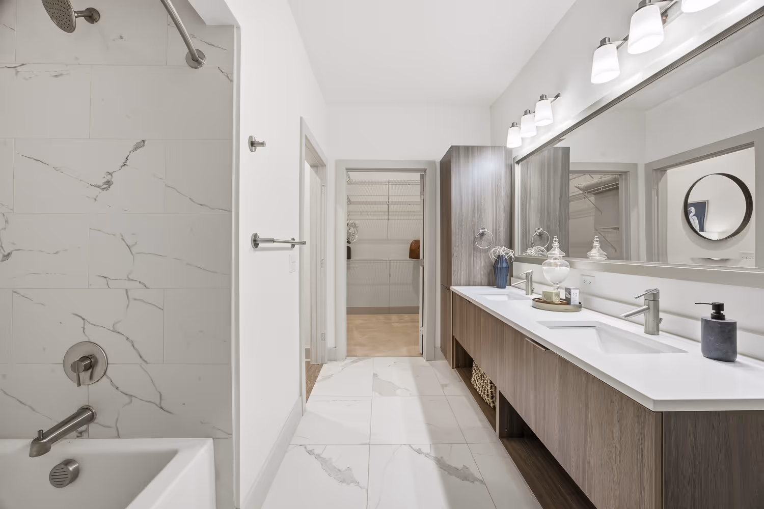 Modern bathroom with white marble floor and wall tiles, double vanity with wood cabinets, large mirror, and walk-in closet visible through doorway.