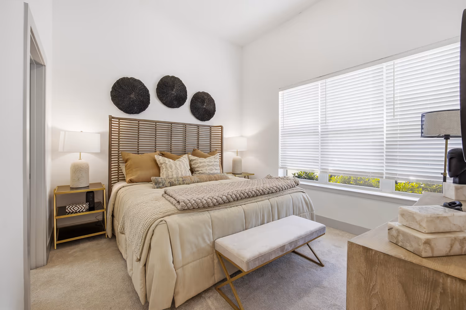 Bright bedroom with beige bedding, rattan headboard, decorative pillows, two bedside lamps, and a large window with white blinds.