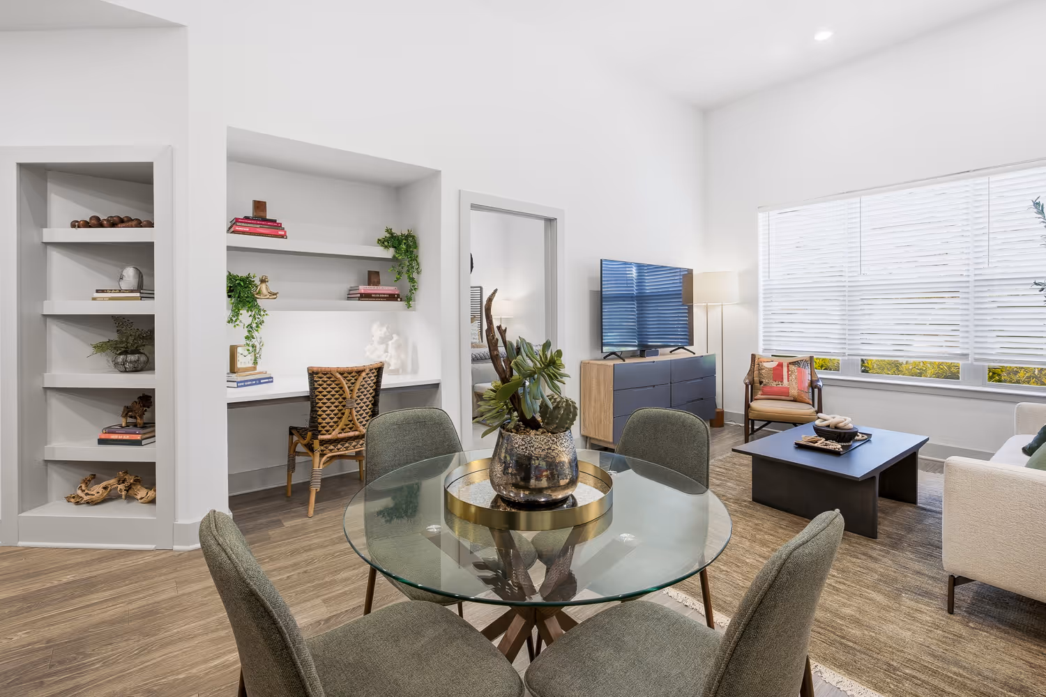 Modern living room with glass dining table, gray upholstered chairs, built-in desk with chair, TV on wooden dresser, and large window with white blinds.