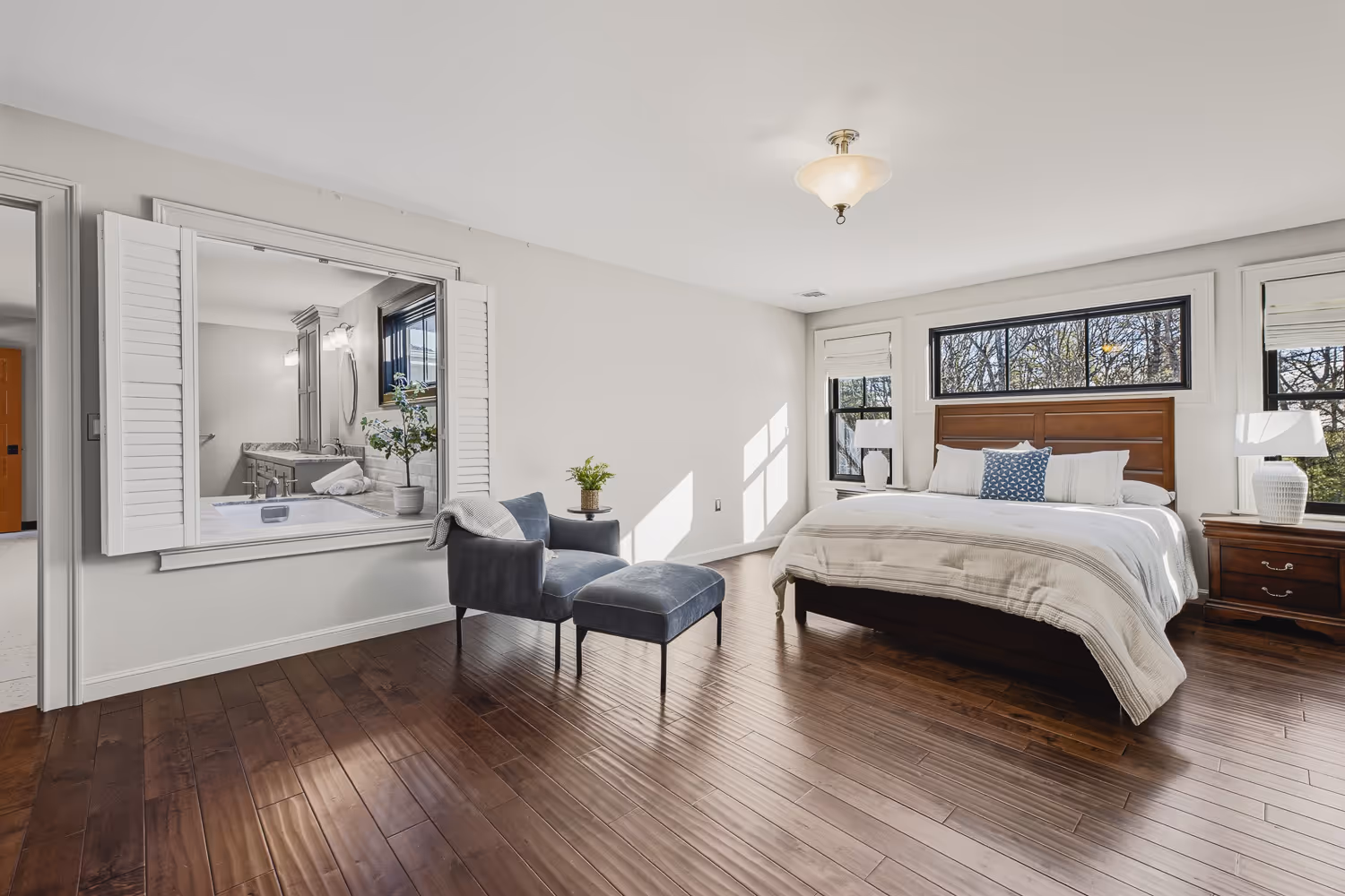 Bright bedroom with wooden floor, king-sized bed with white bedding, blue armchair and ottoman, and window opening into bathroom.