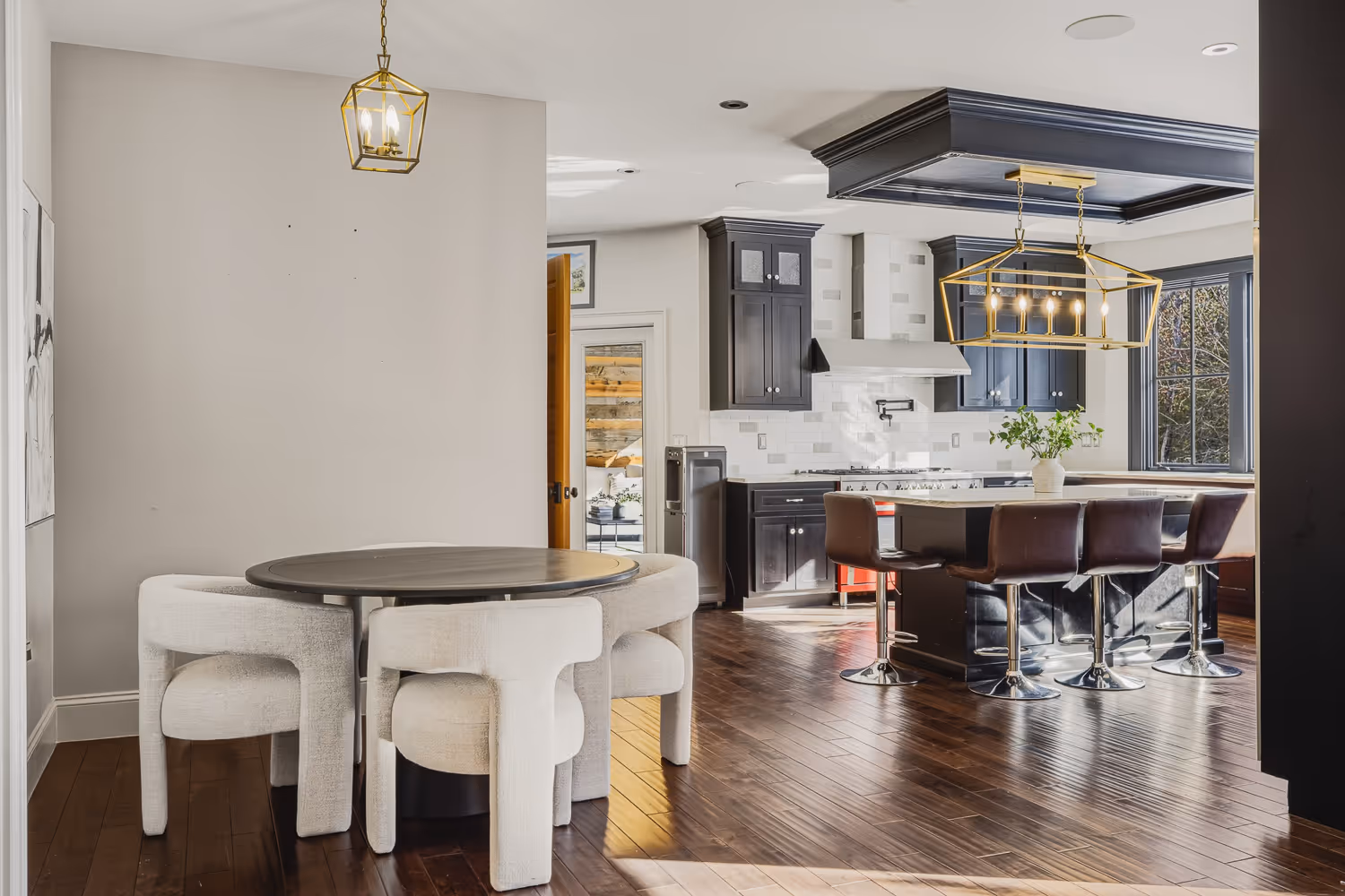Modern kitchen with dark cabinets, an island with brown barstools, white dining table with upholstered chairs, and gold pendant lights.