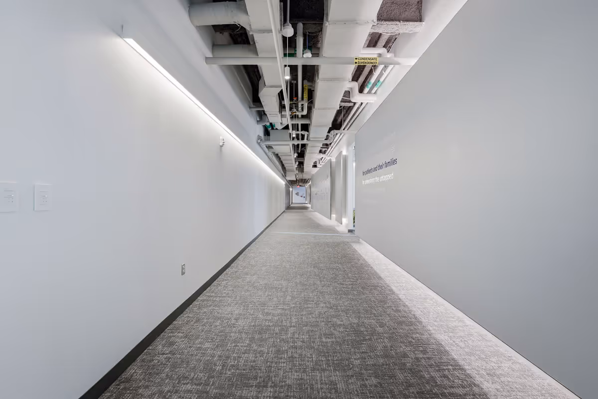 Long, narrow modern hallway with gray carpet, white walls, and exposed ceiling pipes.