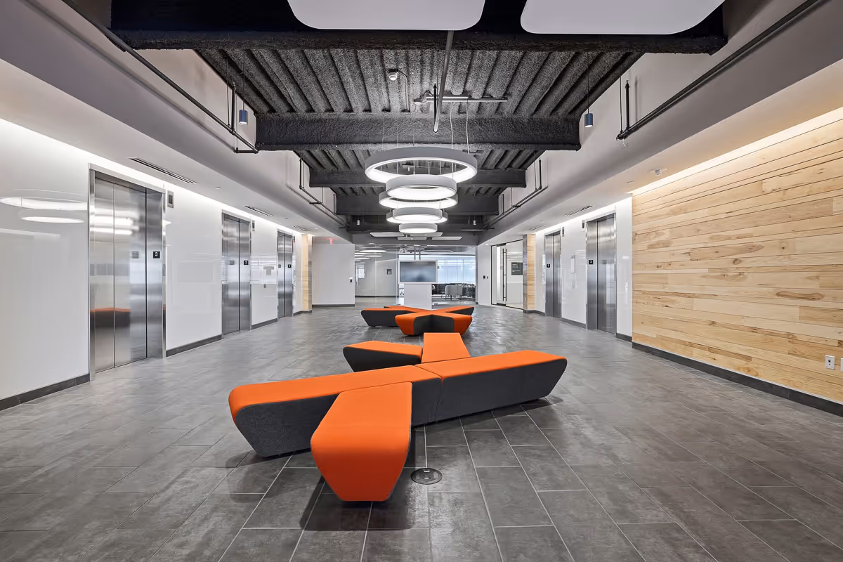 Modern commercial lobby with gray tile flooring, six stainless steel elevators, an orange and black modular bench, and circular ceiling lights.