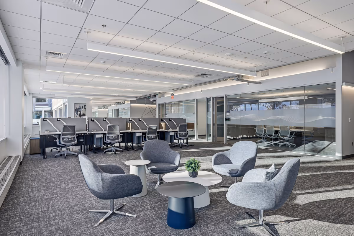 Modern office space with gray swivel chairs arranged around small white and black tables, and cubicles with office chairs in the background.