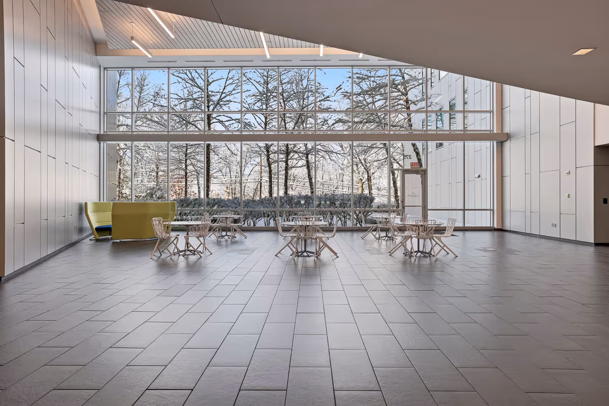 Spacious modern lobby area with white tiled floor, glass tables with chairs, yellow sofa, and large floor-to-ceiling windows showing leafless trees outside.