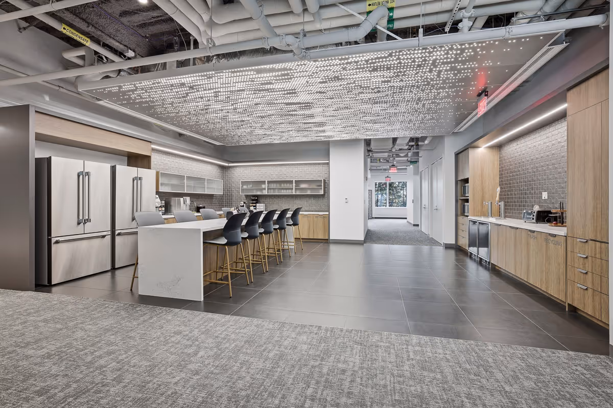 Modern office kitchen with a long island, black high chairs, stainless steel refrigerators, wooden cabinets, and a ceiling panel with numerous small LED lights.