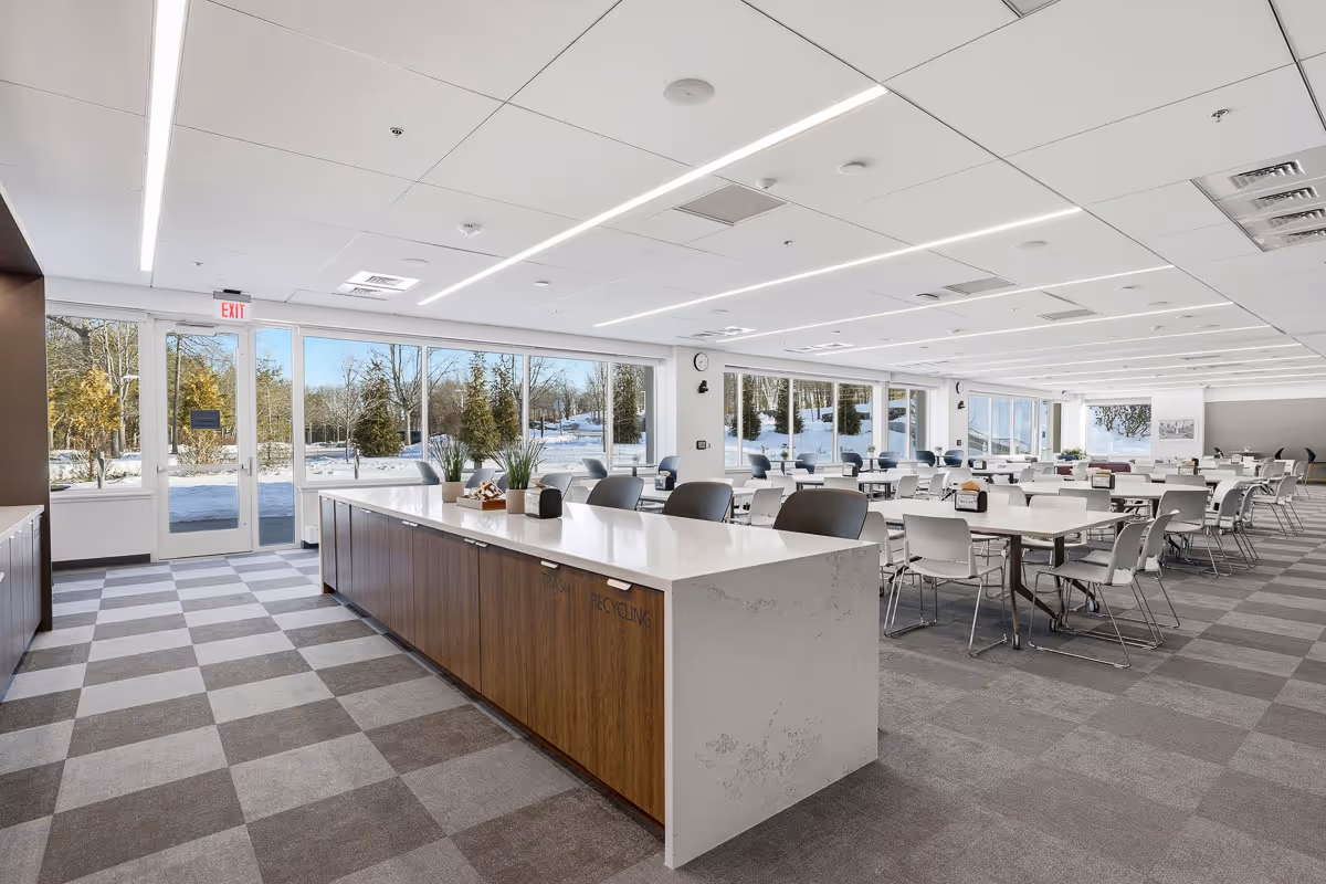 Bright, spacious office dining area with large windows showing snowy outdoor landscape, a long island counter, and multiple tables with chairs.