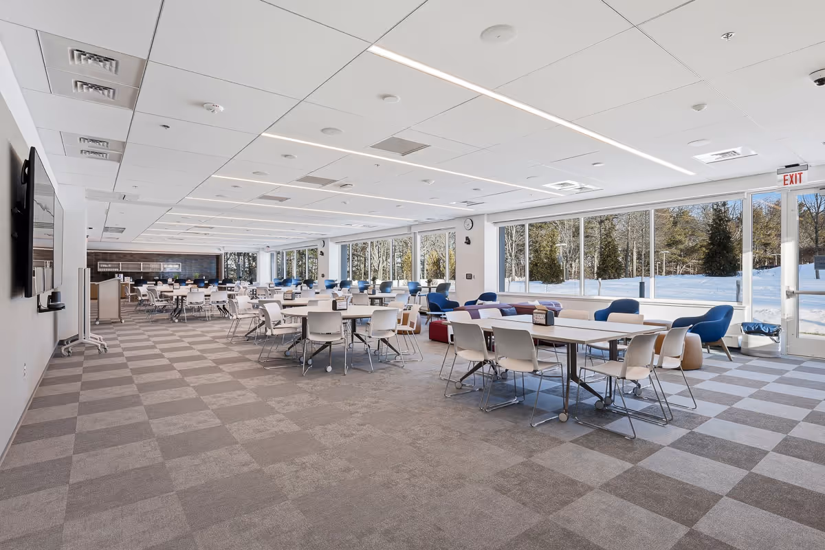 Large modern conference room with multiple white tables, chairs, blue lounge chairs, and large windows showing snowy trees outside.