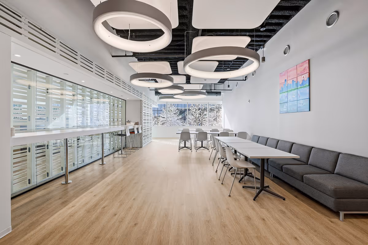 Modern office lounge with circular ceiling lights, long tables, gray chairs, and a gray sofa along the wall under a colorful cityscape painting.