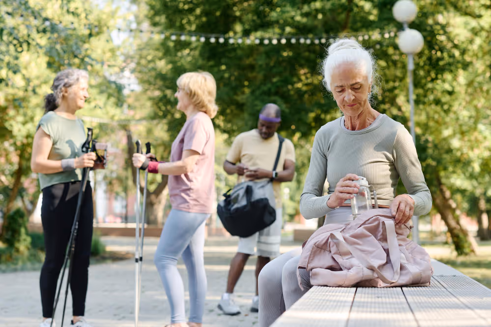 Elderly woman with a walking stick and a bag, standing on a sidewalk, looking ahead with a gentle smile.