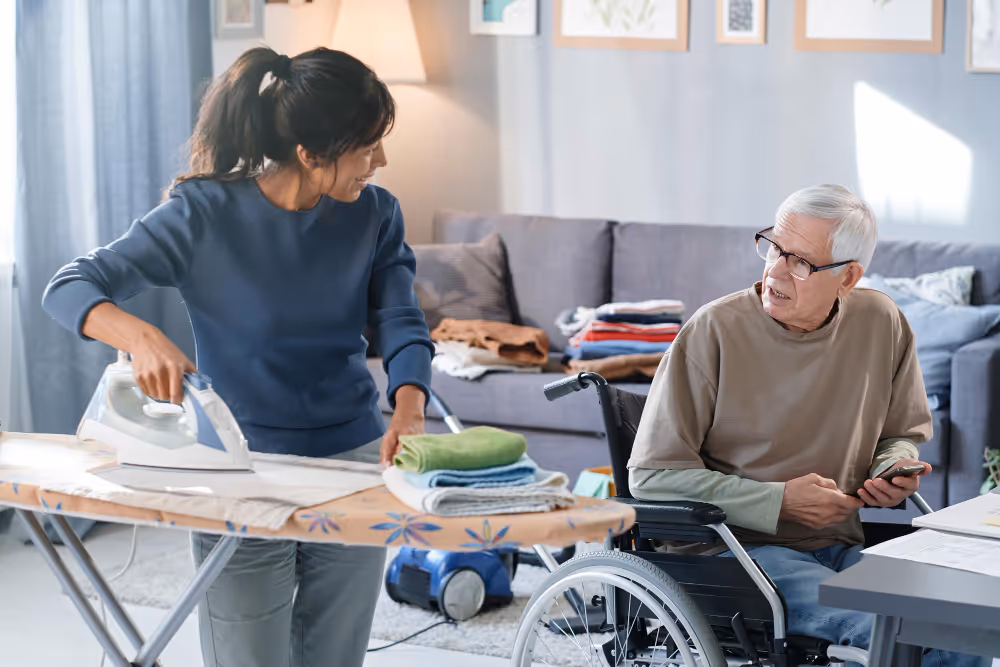 A woman in a wheelchair is ironing clothes alongside an older man, both focused on their task together.