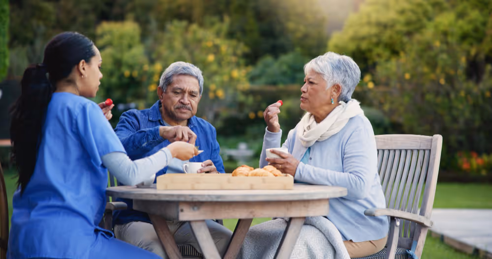 Senior couple enjoying lunch together at an outdoor table, surrounded by greenery and a sunny atmosphere.