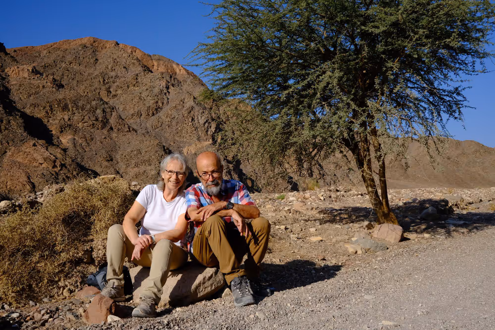 A man and woman sit on a rock in a vast desert landscape, surrounded by sandy terrain and clear blue skies.