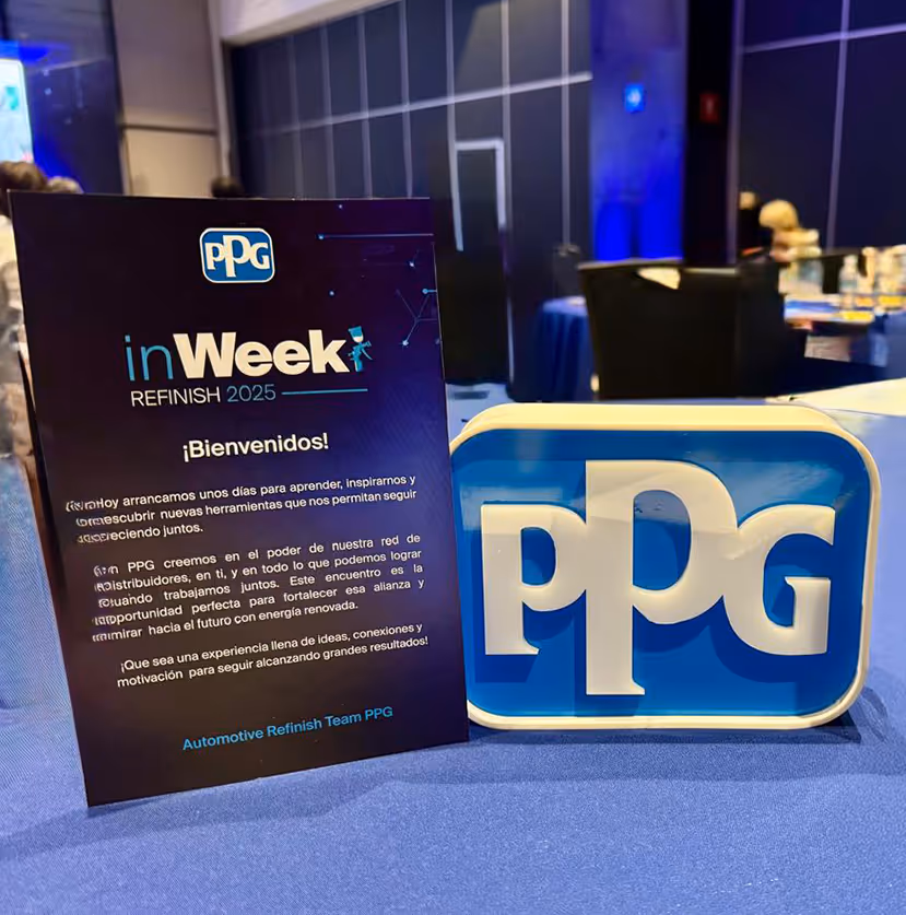 Cartel en mesa con el logotipo de PPG y texto de bienvenida al evento inWeek Refinish 2025 en un salón de conferencias.