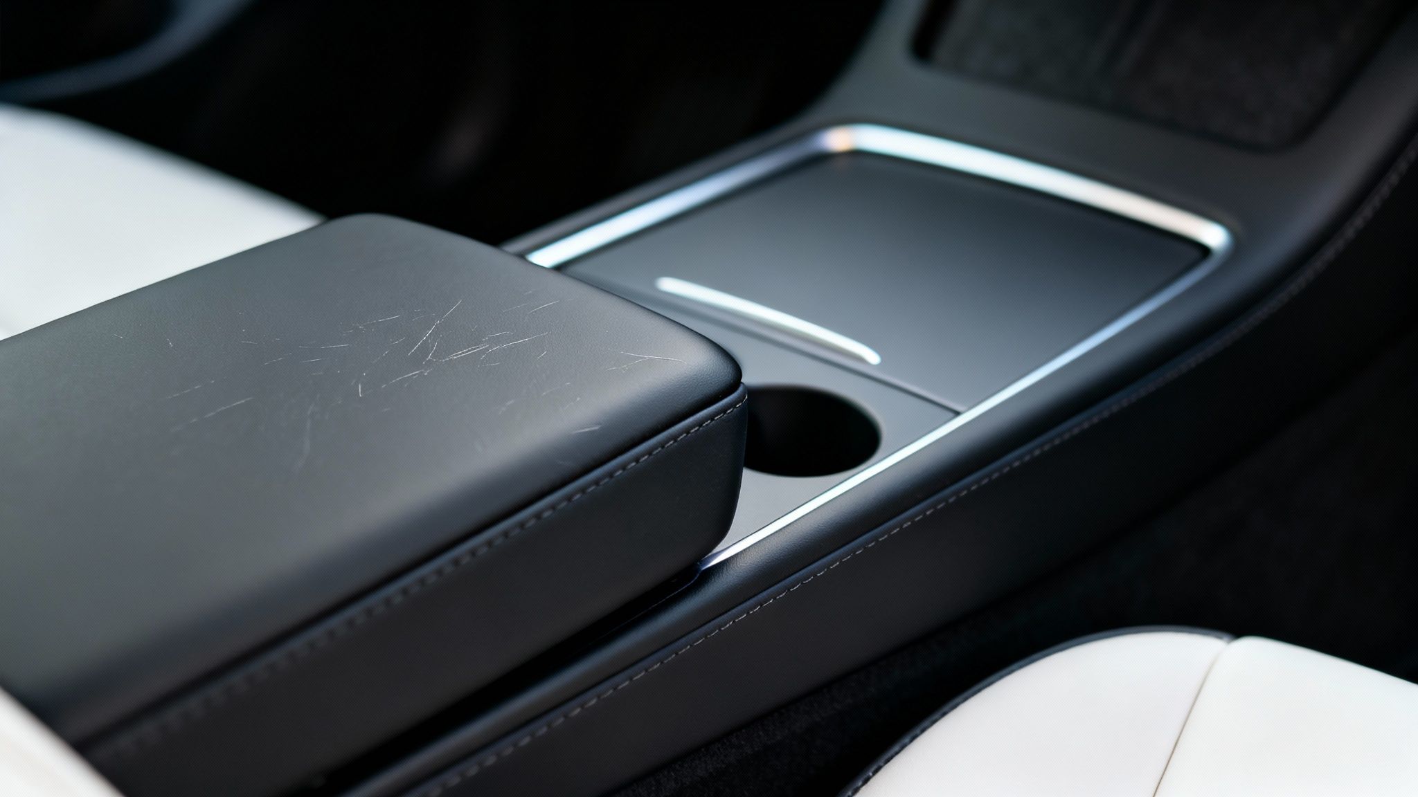 Close-up of a scratched black car armrest and center console with white seats.
