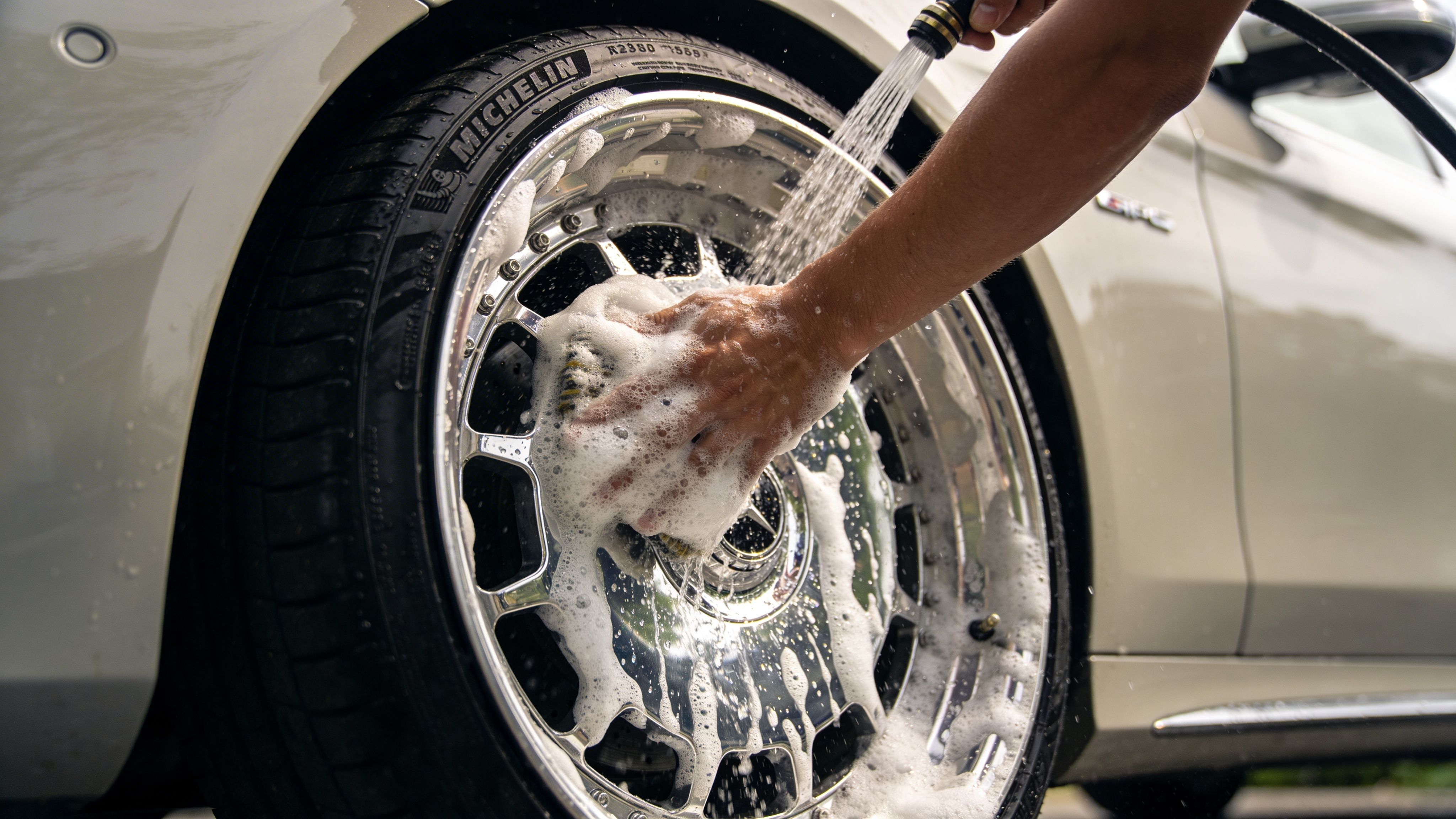 A person washing a shiny car wheel with soap and water using a sponge and hose.