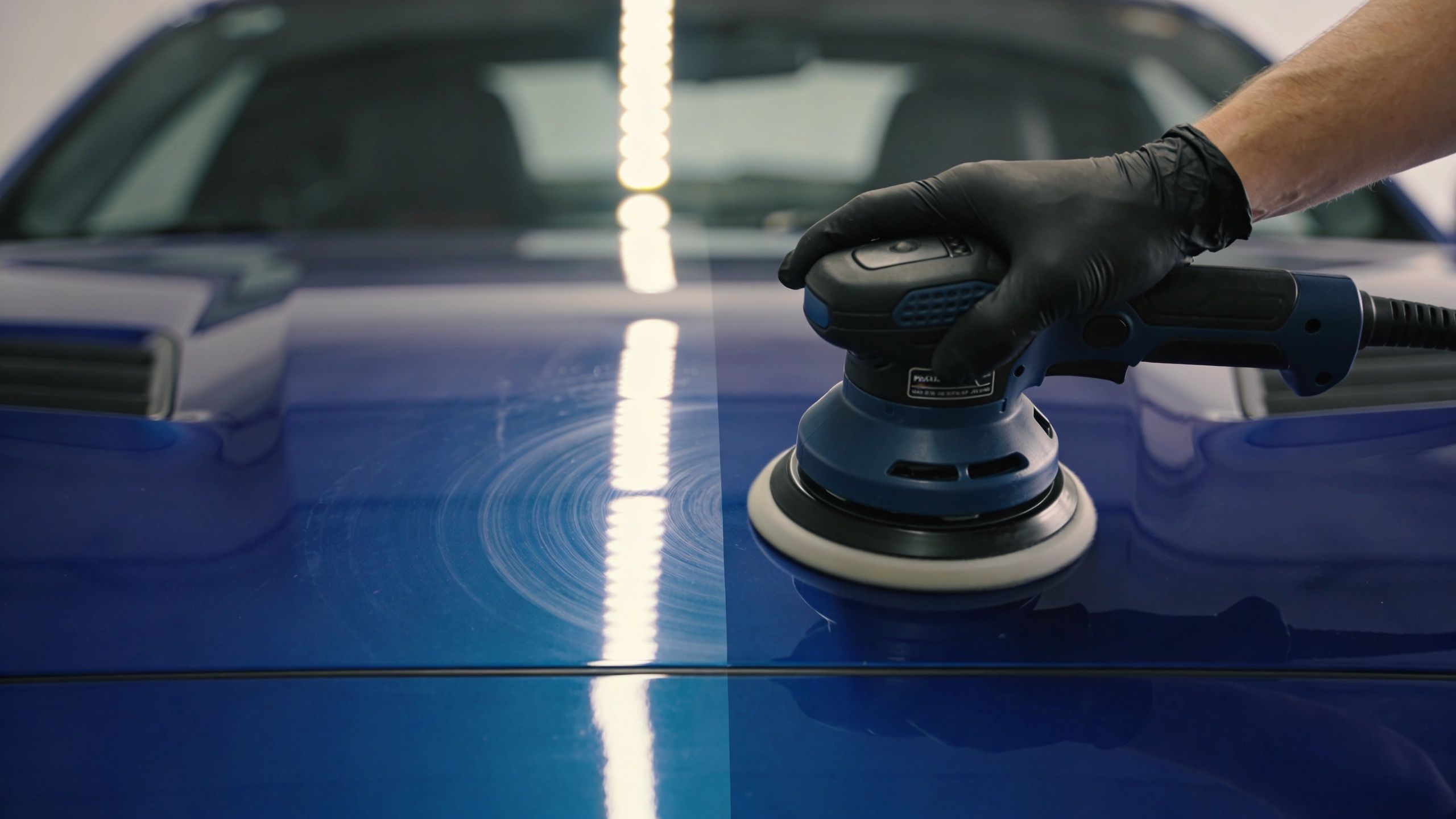 A professional technician uses an orbital polisher to perform paint correction on a shiny blue car hood.