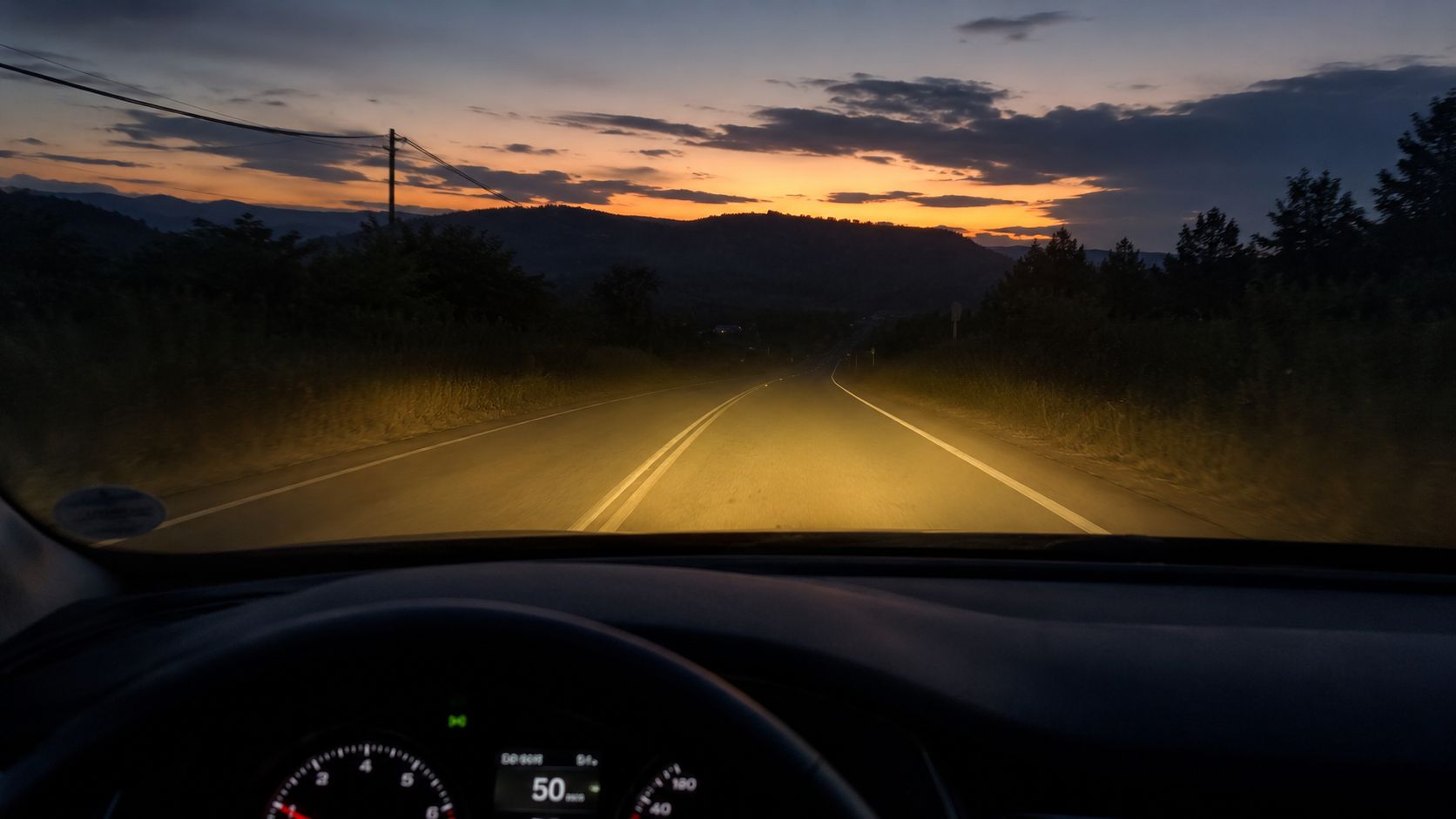 A view from inside a car driving on a road at dusk with illuminated headlights.