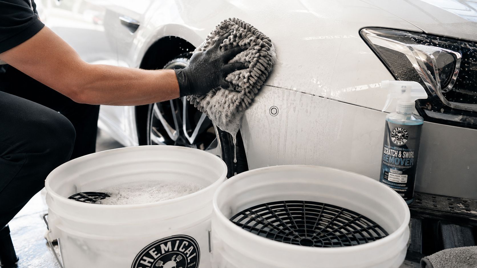 A professional detailer using a microfiber wash mitt to clean a white car with scratch remover spray.