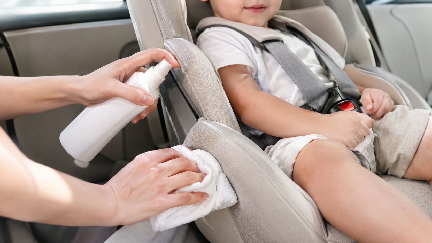 A caregiver cleaning a toddler's car seat with a spray bottle and a soft white cloth.