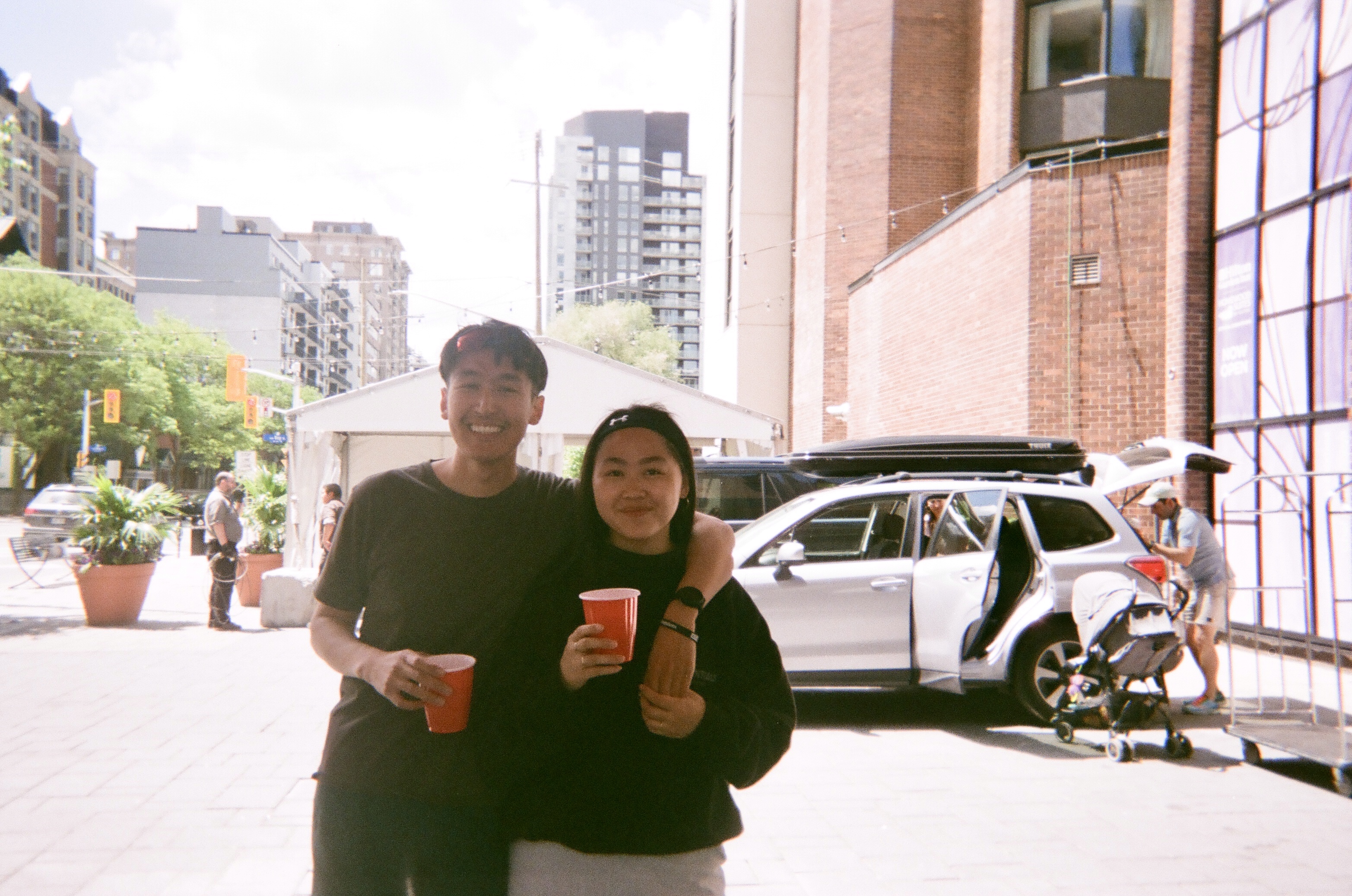 A smiling young man and woman holding red cups stand side by side outdoors with city buildings and a white car in the background.