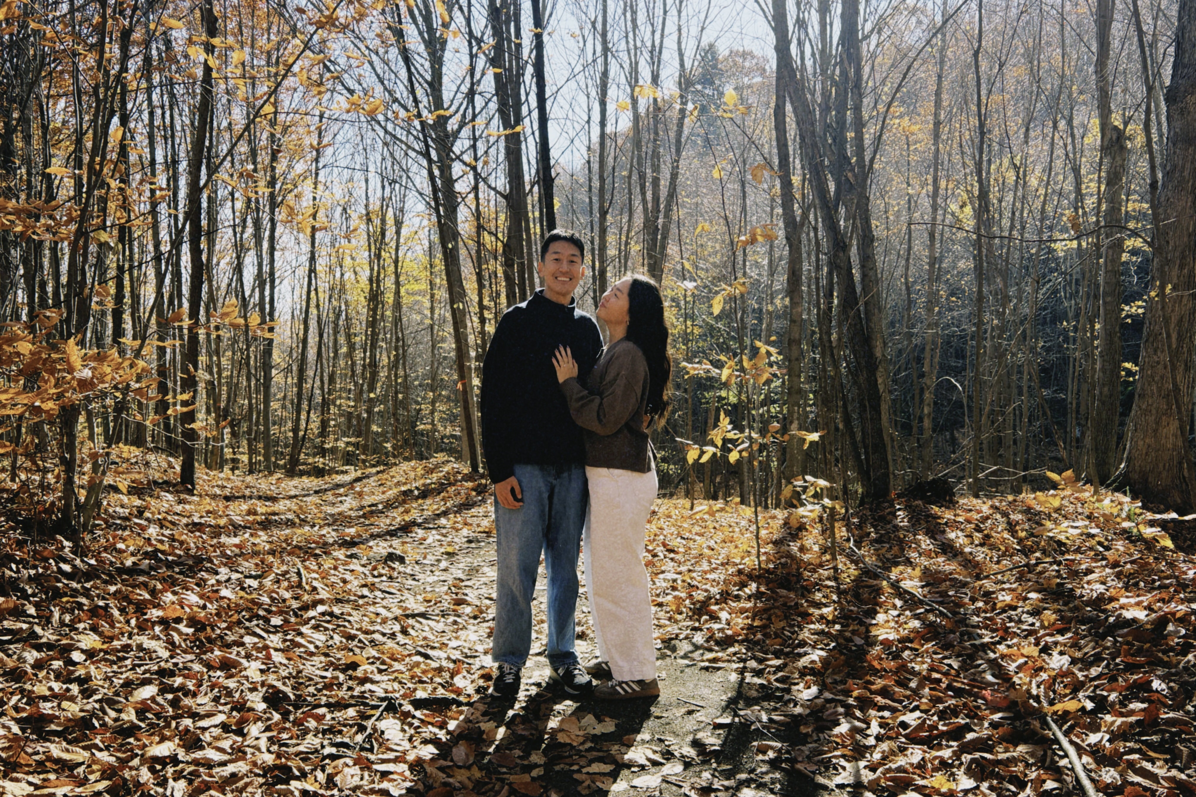 Couple standing close together on a leaf-covered forest path in autumn with sunlight casting long shadows.