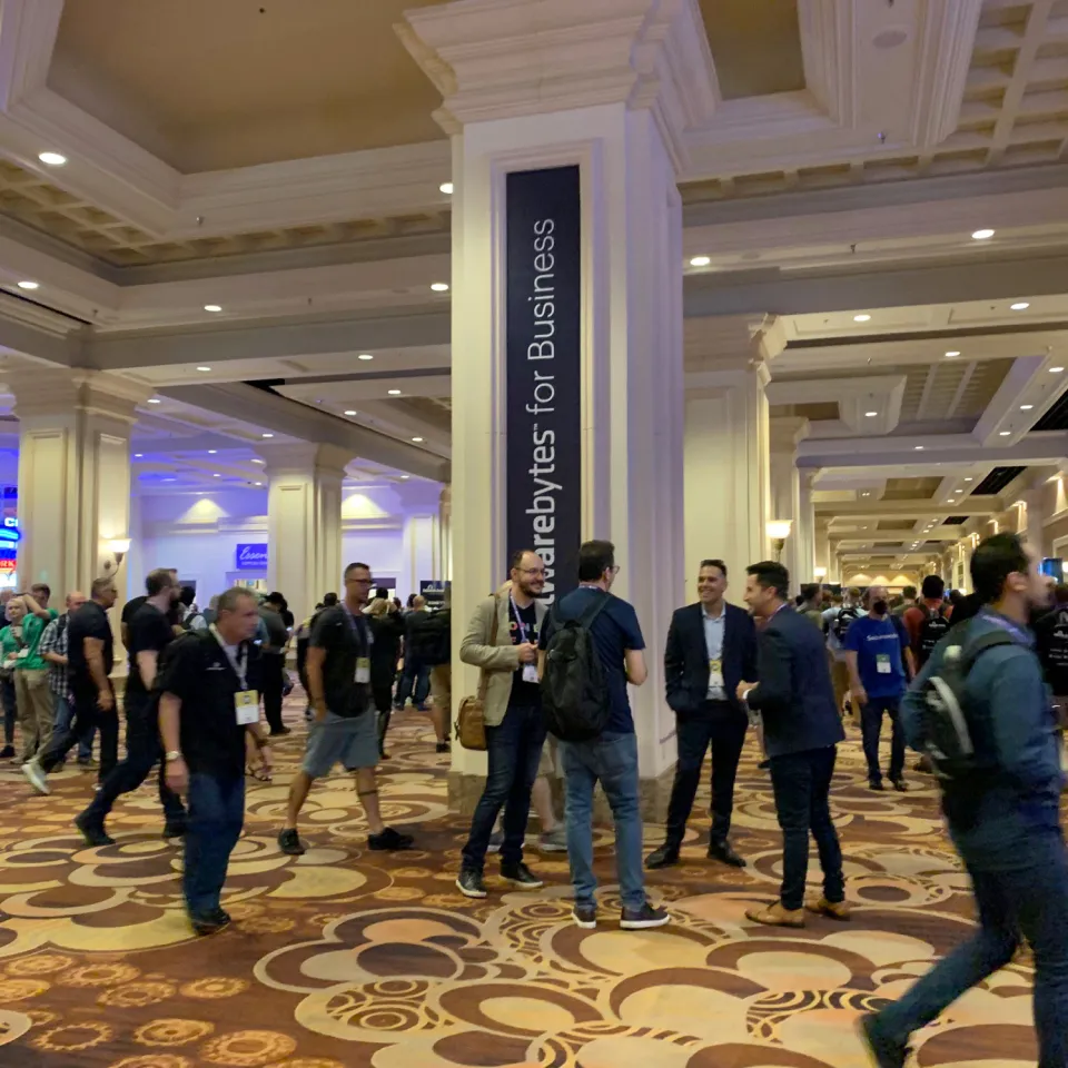 Businessmen gather in front of a 'Malwarebytes for Business' ad on a column in an event center.
