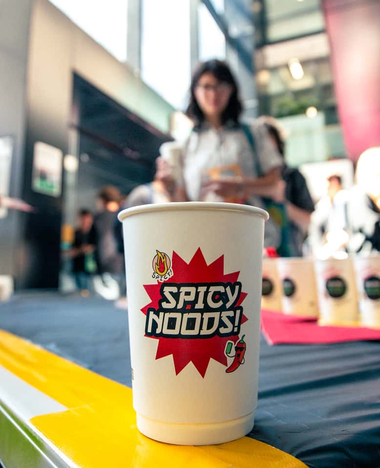 Closeup photo of a Spicy Noods branded cup of instant noodles on a table with people in the background