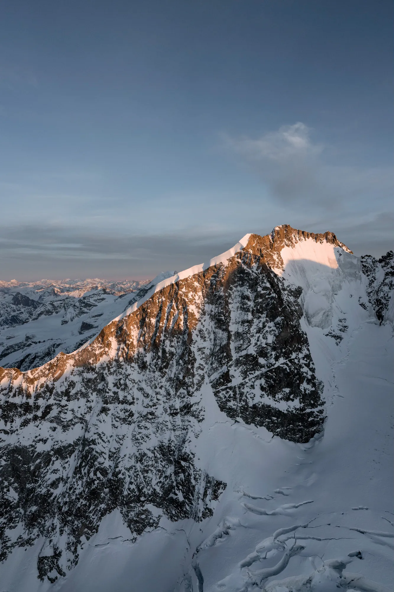 Schneebedeckter Berggipfel im Abendlicht mit weitläufigen Alpen im Hintergrund unter blauem Himmel.