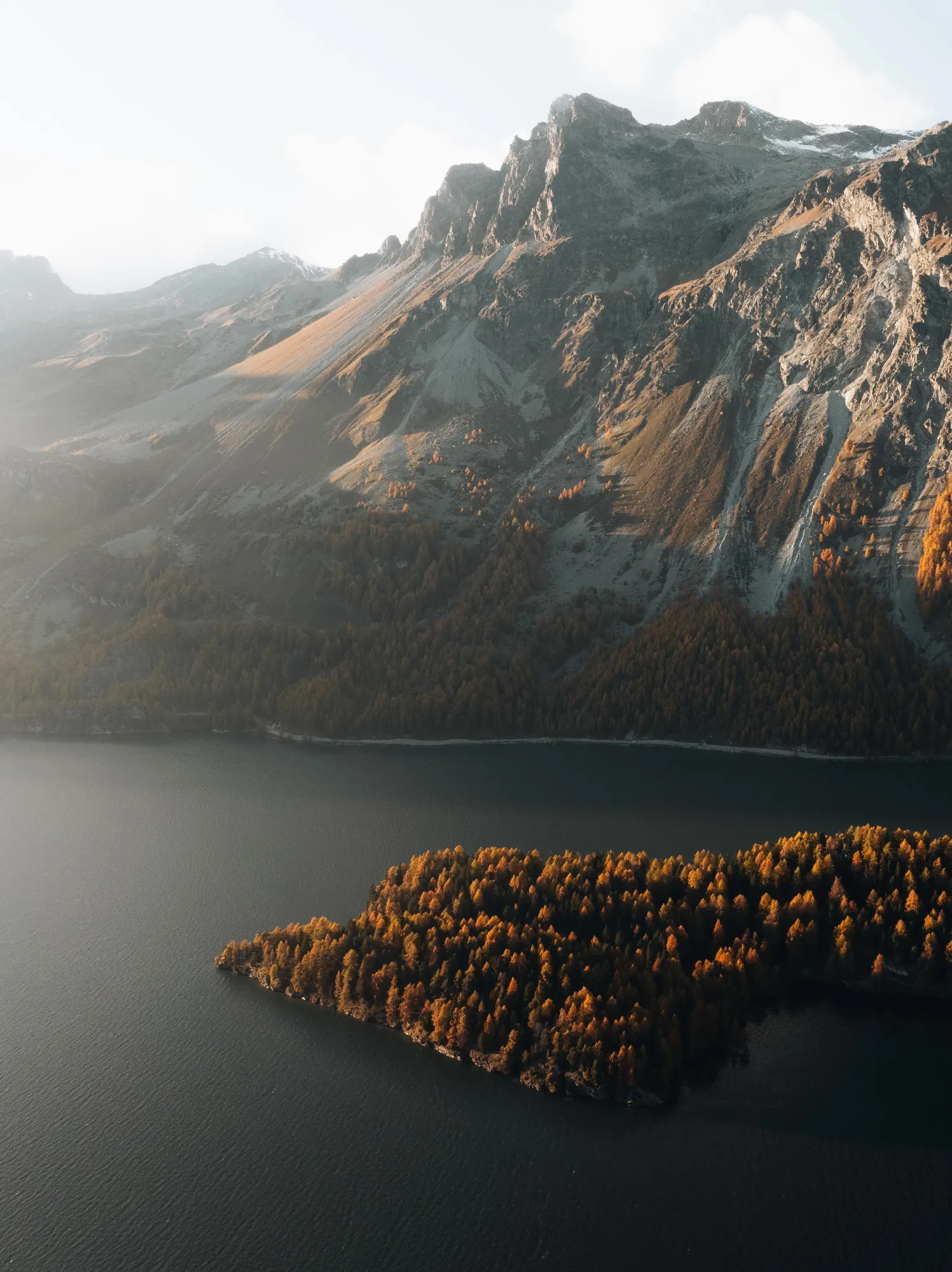 Berglandschaft mit herbstlich gefärbtem Wald an einer Halbinsel und einem ruhigen See im Vordergrund.