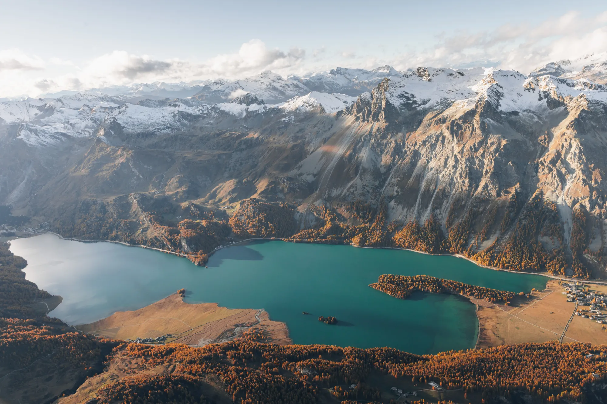 Herbstliche Berglandschaft mit blauem See, goldenen Wäldern und schneebedeckten Gipfeln unter einem bewölkten Himmel.