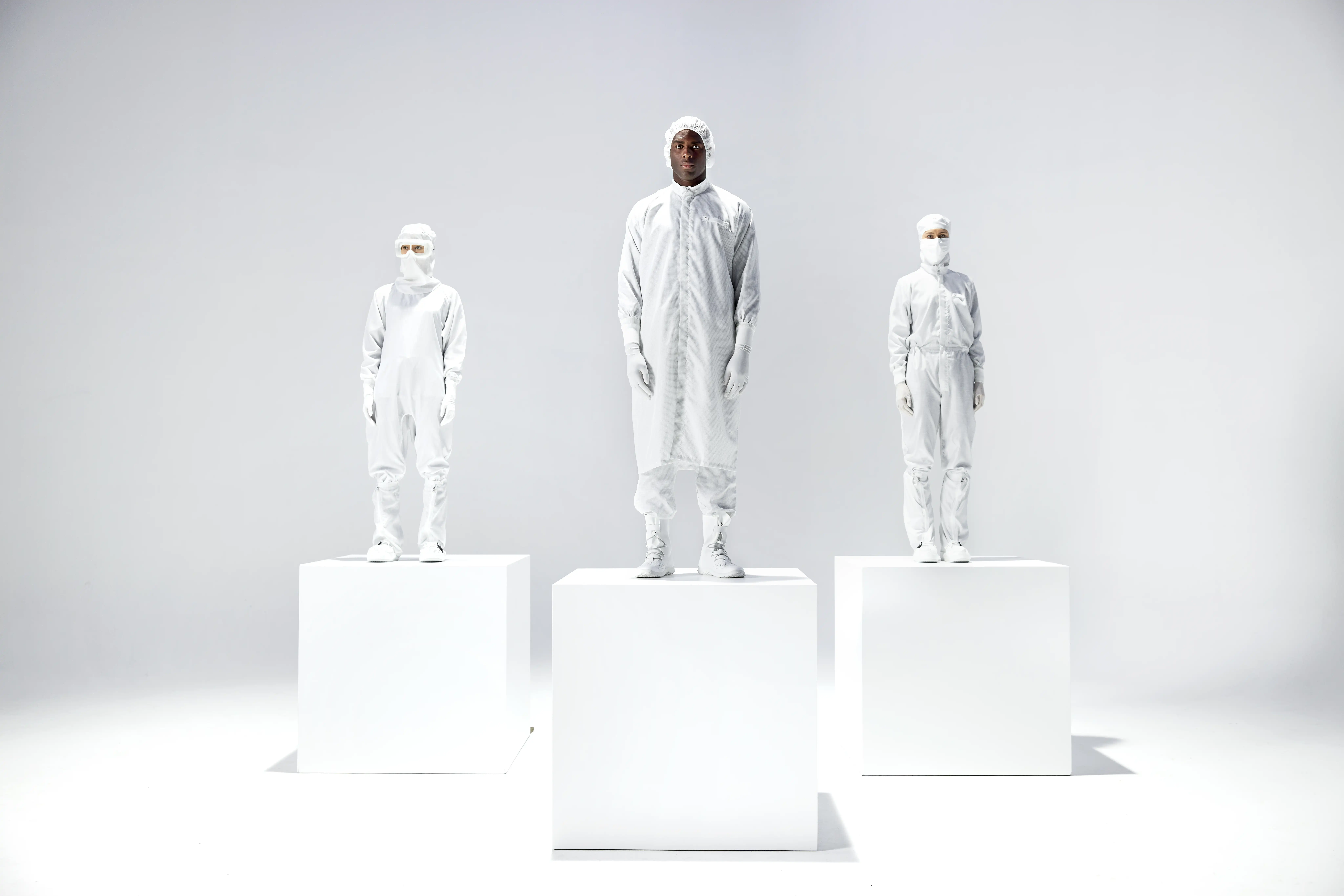 Four figures in white protective suits stand on white pedestals against a light gray background, conveying a modern, minimalist aesthetic.
