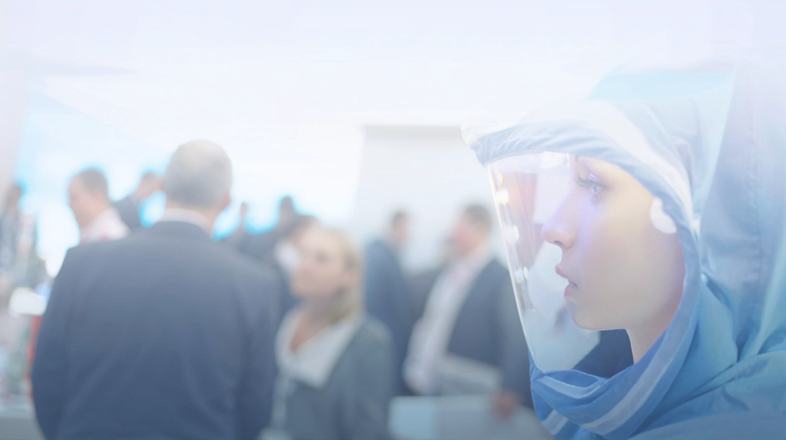 Person mit Schutzanzug und Gesichtsvisier im Vordergrund, unscharfe Menschenmenge im Hintergrund.