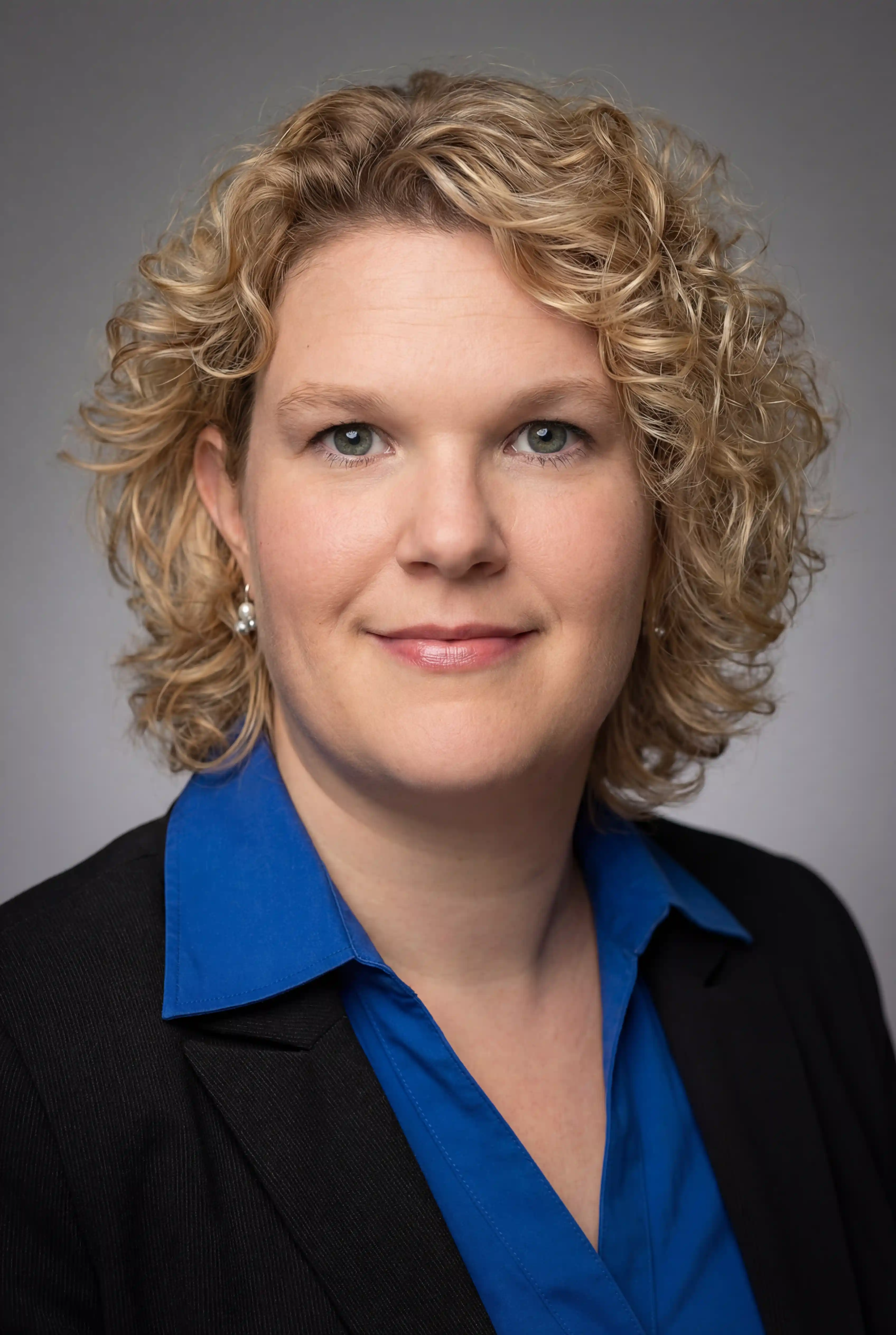 Porträt einer lächelnden Frau mit lockigem blondem Haar, die eine blaue Bluse und ein schwarzes Sakko trägt.