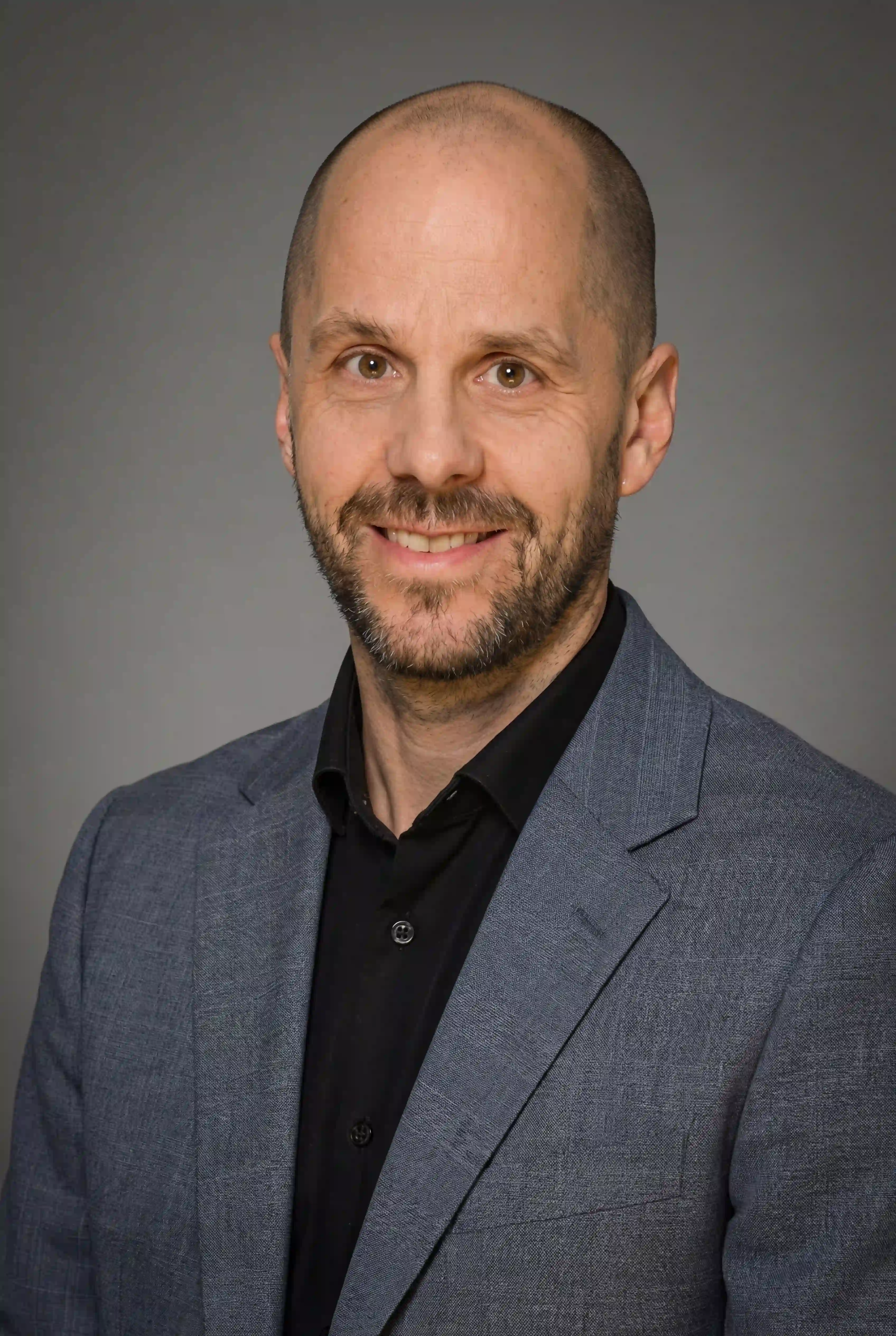 Porträt eines lächelnden Mannes mit Glatze und Bart, der einen grauen Blazer und ein schwarzes Hemd trägt.