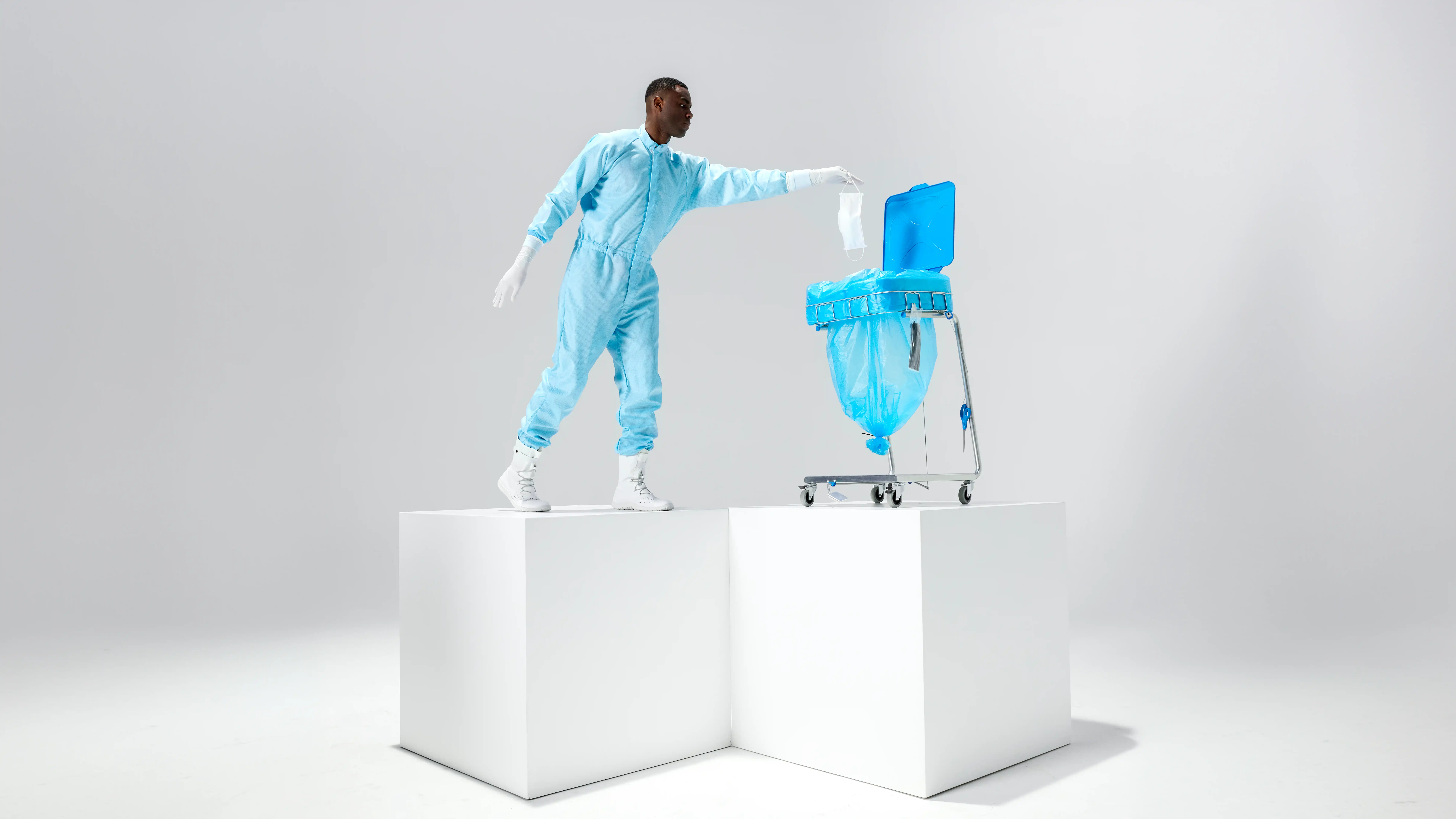 Eine Person im blauen Schutzanzug wirft eine medizinische Maske in einen blauen Müllsack auf einem Metallwagen.